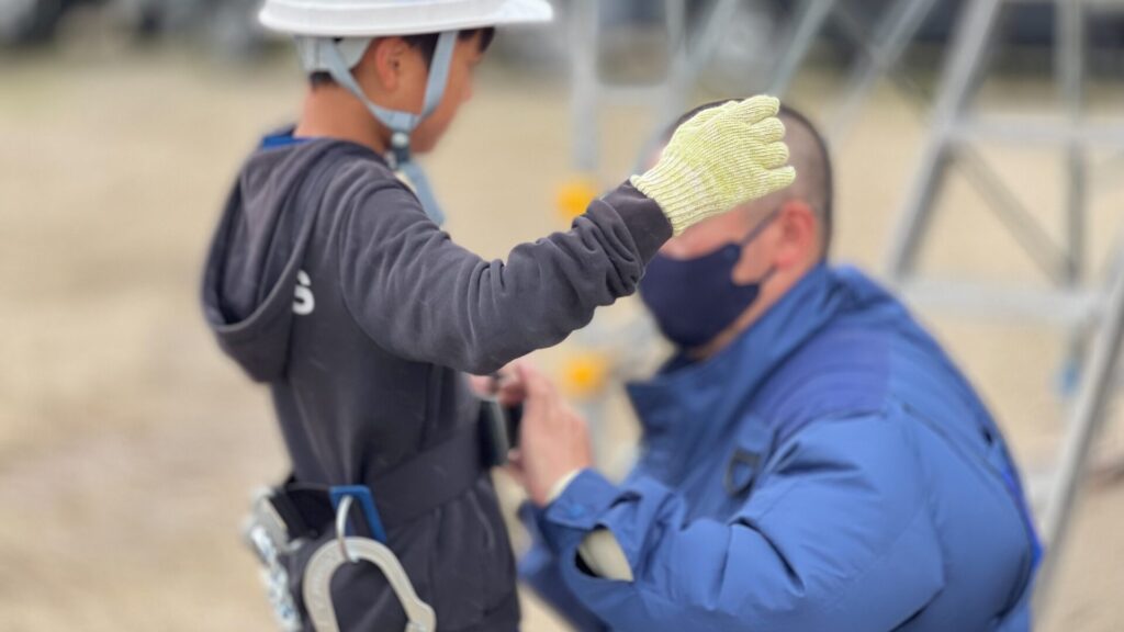 ヘルメットと軍手を装着した子どもが、足場組み立て作業のサポートや説明を受けている様子