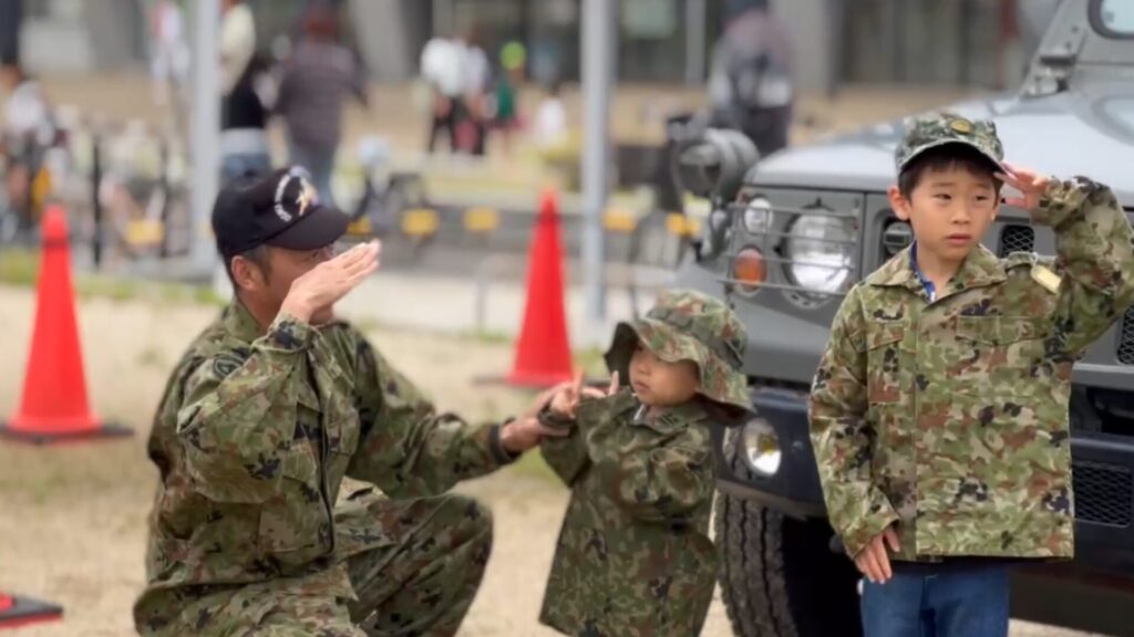 自衛隊員に敬礼のポーズを教わりながら、迷彩服姿で敬礼する子どもたちの様子